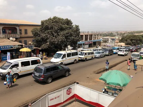 Minibuses, an SUV and cars queueing on a busy road in Kumasi, with roadside stalls and a large shade tree