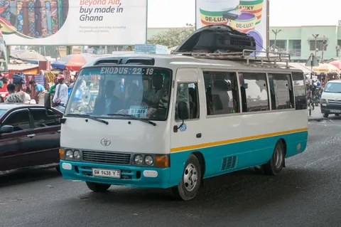 A white and blue Toyota Coaster minibus driving on a busy street in Accra, Ghana