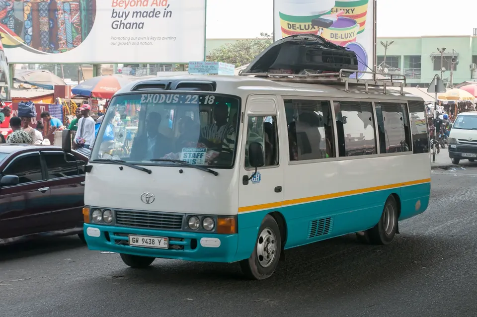 A white and blue Toyota Coaster minibus driving on a busy street in Accra, Ghana