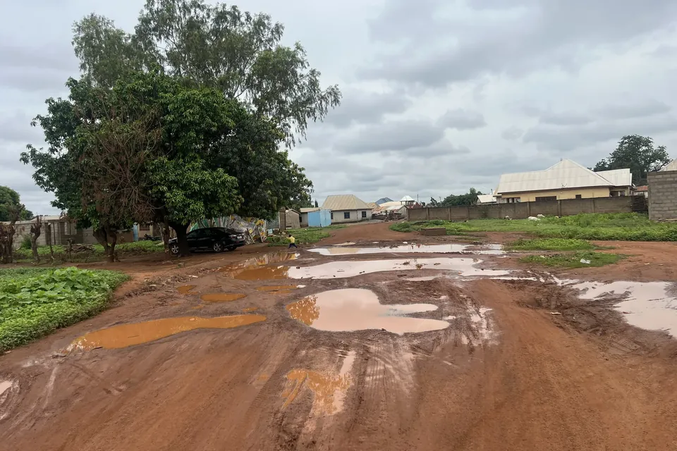 A red-earth unpaved road in Northern Ghana with rainwater-filled potholes after rain, a parked car and houses beyond